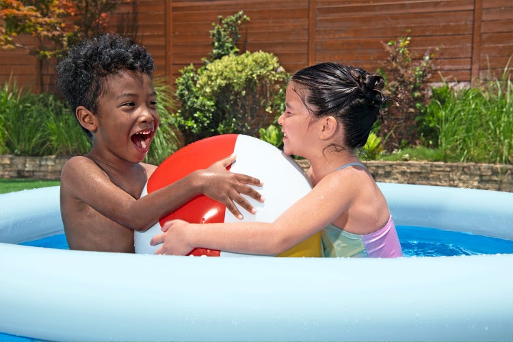 Zwei Kinder spielen mit einem Wasserball in einem hellblauen aufblasbaren Pool in einem sonnigen Garten.
