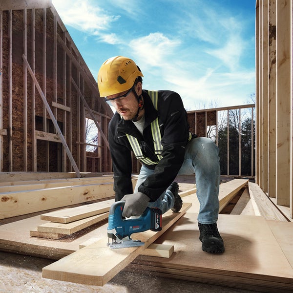 Ein Bauarbeiter mit Schutzhelm und Arbeitskleidung sägt auf einer Baustelle mit einer Bosch Akku Stichmaschine ein Holzbrett zu.