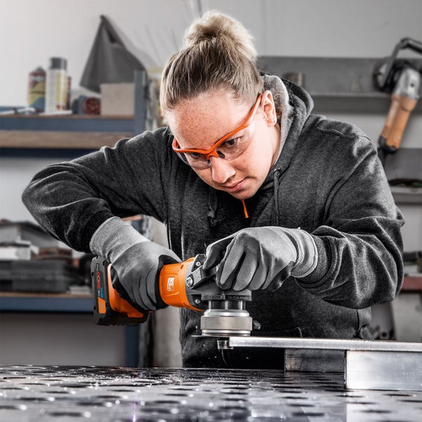 Eine Frau bearbeitet Metall mit einer Fein Akku-Kantenfräse in einer Werkstatt, sie trägt Schutzbrille und Arbeitshandschuhe.