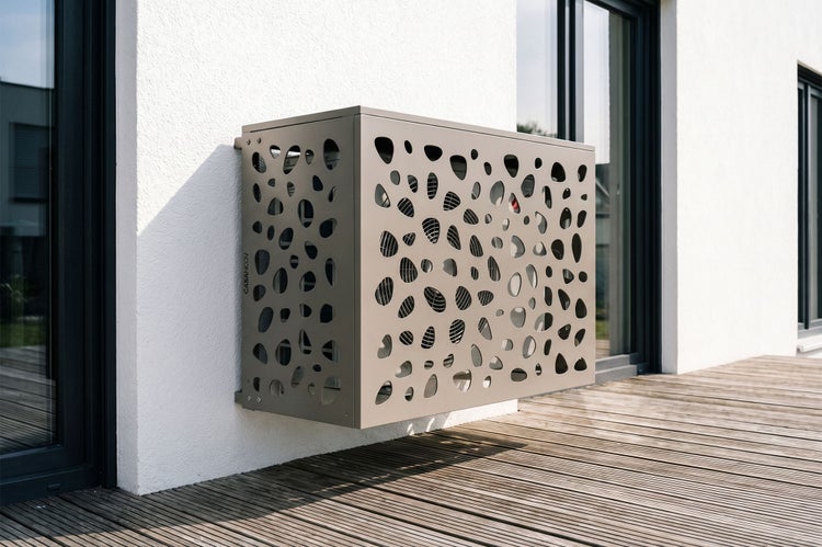 Wandmontierte Klimaanlagen-Verkleidung aus Metall mit organischem Lochmuster an einer Hauswand über einer Holzterrasse.