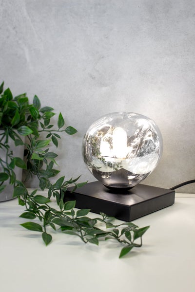 Moderne Tischleuchte mit gläserner Kugel auf einem schwarzen Sockel neben einer Pflanze.
