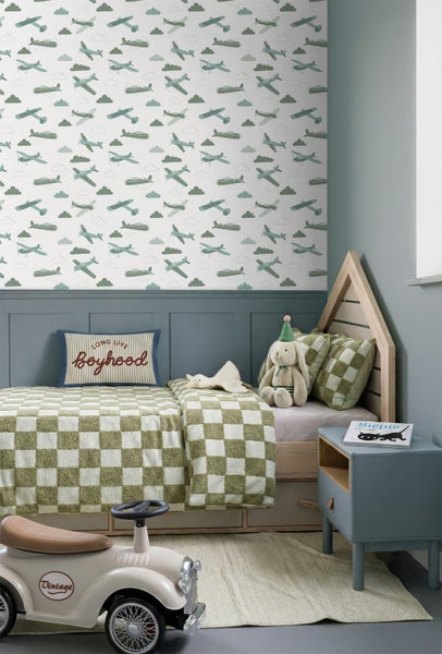 Kinderzimmer mit Flugzeugtapete, hölzernem Hausbett, karierter Bettwäsche, blauem Nachttisch und einem Vintage Tretauto auf einem Teppich.