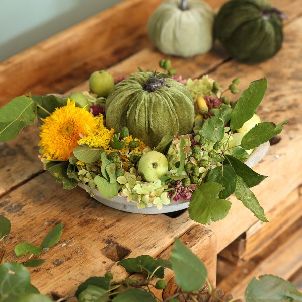 Herbstliche Tischdekoration in einer Schale mit Stoffkürbis, Sonnenblume, Äpfeln und herbstlichen Pflanzen auf einer hölzernen Unterlage.