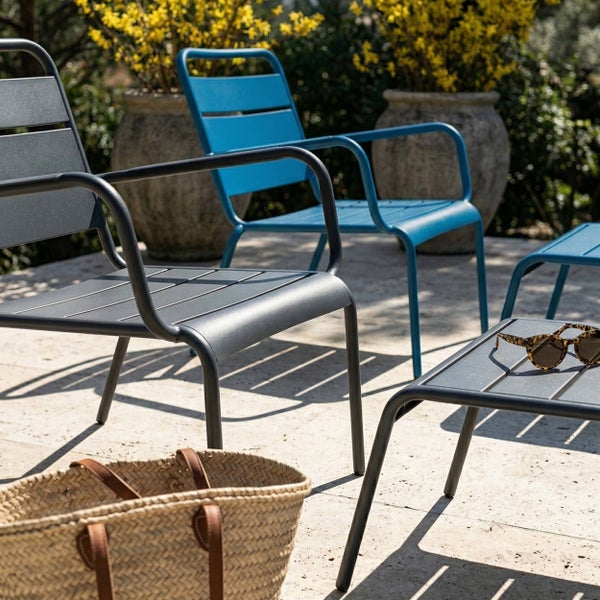 Moderne Gartenstühle und Beistelltische aus Metall in Grau und Blau auf einer hellen Steinterrasse mit einem Weidenkorb und Pflanzen.