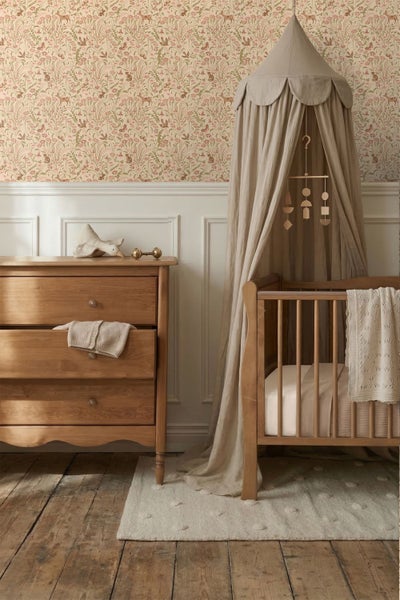 Kinderzimmer mit Holzkommode und Babybett vor einer Wand mit geblümter Tapete und weißer Wandvertäfelung. Ein Stoffhimmel hängt über dem Bett.