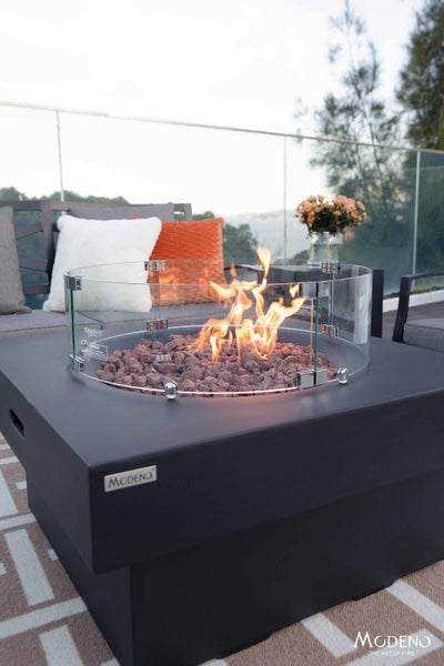 Modeno Gasfeuerstelle mit quadratischem schwarzem Korpus, Lavasteinen und Glaswindschutz auf einer Terrasse mit Gartenmöbeln.