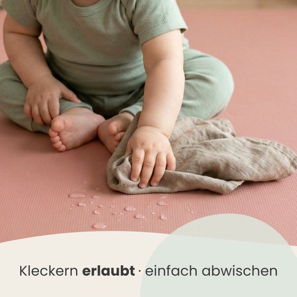 Ein Baby wischt Flüssigkeit von einer strukturierten Spielmatte mit einem Tuch ab.
