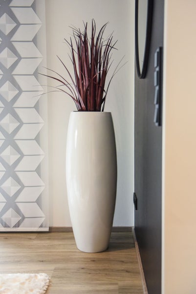 Hohe weiße Bodenvase mit dunkelrotem Dekogras in einem modernen Raum mit Holzfußboden und geometrischem Wandmuster.