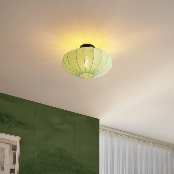 Eingeschaltete Deckenleuchte mit ovalem, geripptem Stoffschirm an einer weißen Decke in einem hellen Zimmer mit grüner Wand und Vorhang.