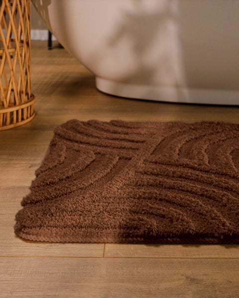 Braune, strukturierte Badematte mit Wellenmuster auf Holzboden vor einer weißen Badewanne.