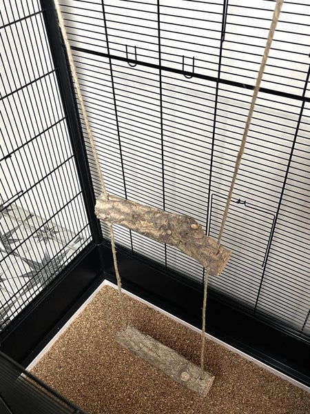 Zweistöckige Vogelschaukel aus Naturholz und Seil in einem Vogelkäfig mit Einstreu am Boden.