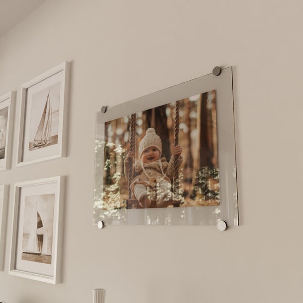 Wandgestaltung mit Acrylglasfoto eines Kindes auf einer Schaukel sowie gerahmten Bildern von Segelschiffen an einer hellen Wand.