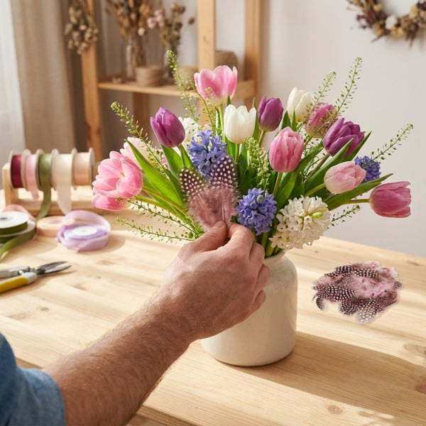 Person arrangiert Frühlingsblumenstrauß mit Tulpen, Hyazinthen und Dekofedern in einer weißen Vase auf einem Holztisch.