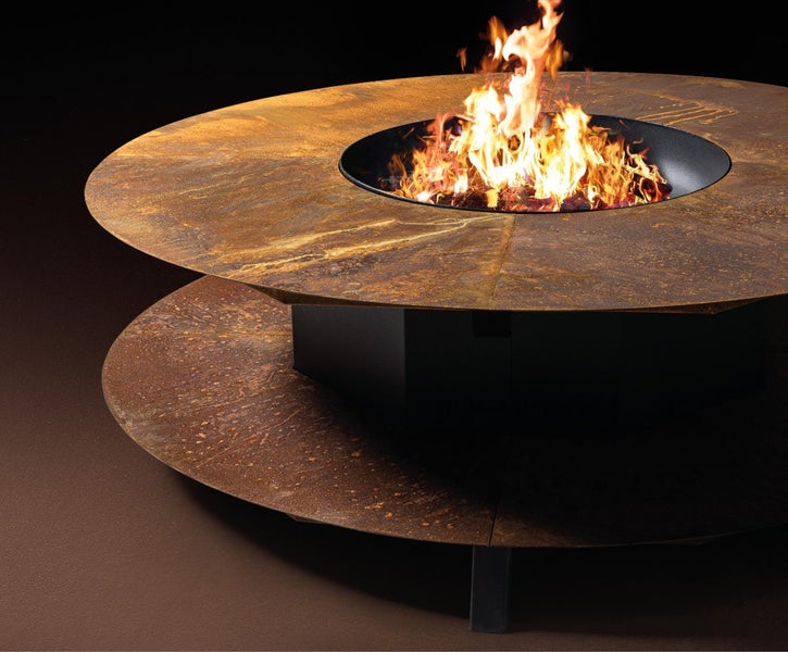 Runde Feuerstelle mit Grillring aus Metall in Edelrostoptik mit brennendem Feuer im Zentrum.