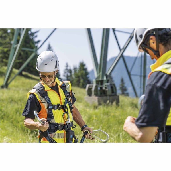 Arbeiter mit Auffanggurt, weißem Schutzhelm und Warnweste hält Karabinerhaken im Freien vor einem Gittermast.
