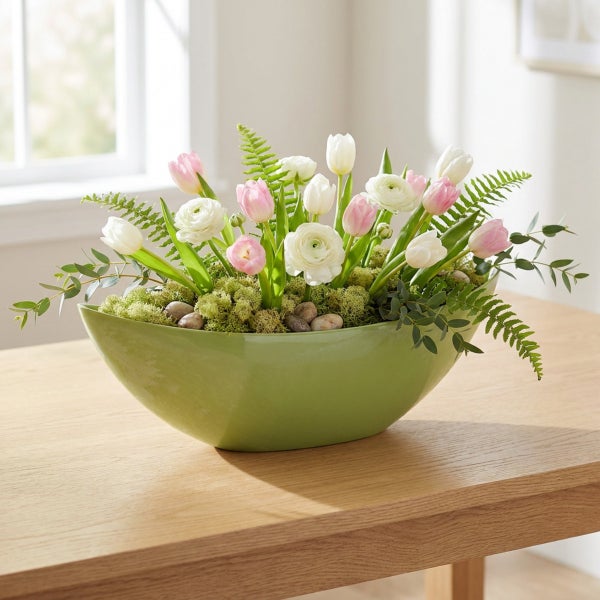 Frühlingshaftes Blumenarrangement mit Tulpen und Ranunkeln in einer ovalen Schale auf einem Holztisch.