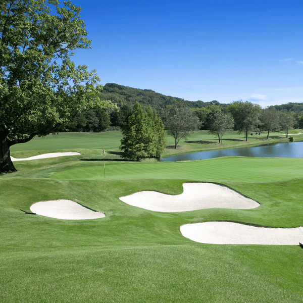 Golfplatz mit gepflegtem Rasen, Sandbunkern, einem Teich und Bäumen unter blauem Himmel.