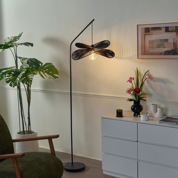 Moderne Stehleuchte mit fächerförmigem Schirm aus dunklem Geflecht in einem hellen Wohnzimmer neben einer Kommode und einer Zimmerpflanze.