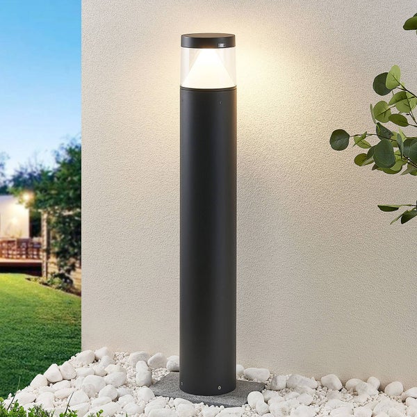 Moderne schwarze LED-Außenstehleuchte in Säulenform vor einer hellen Wand auf weißen Steinen.
