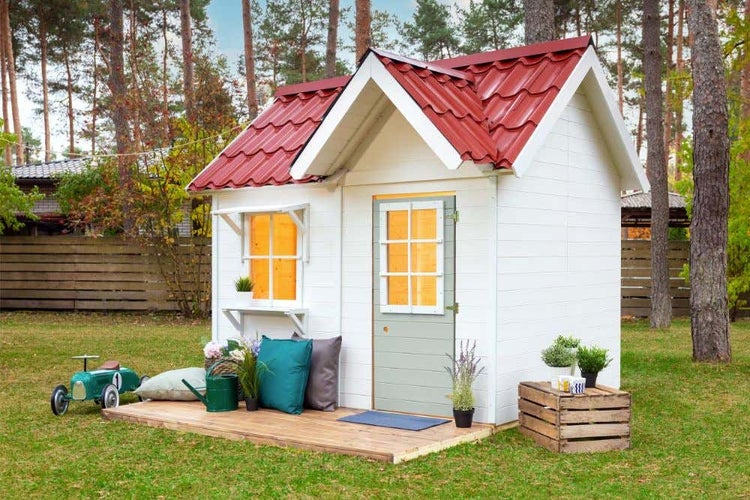 Weißes Kinderspielhaus aus Holz mit rotem Dach, grüner Tür und Fenster im Garten auf einer Holzterrasse mit Spielzeug und Pflanzen.