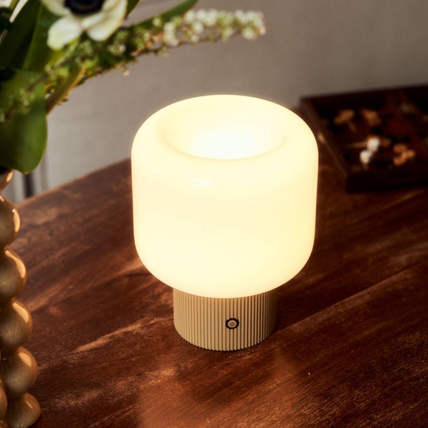 Kleine Tischleuchte mit abgerundetem Schirm und geriffeltem Sockel mit Einschaltknopf, helles warmes Licht auf einem Holztisch neben einer Vase.