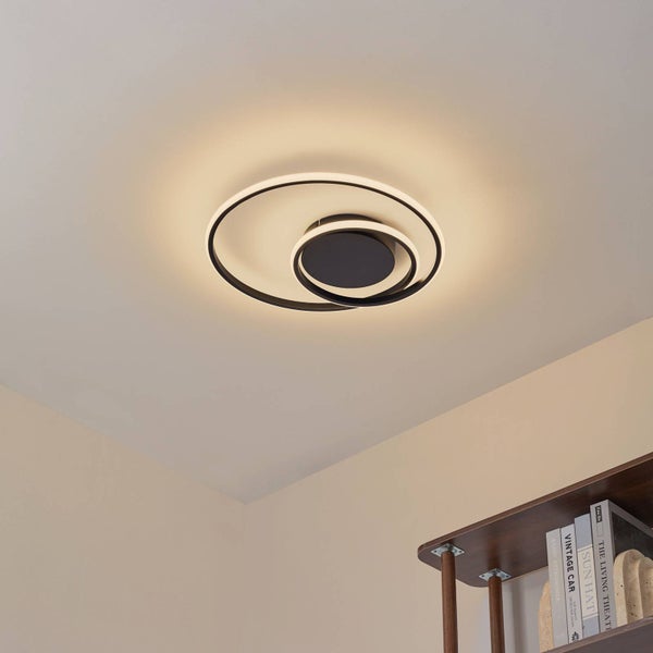 Moderne LED-Deckenleuchte mit ineinander verschlungenen schwarzen Ringen in einem hellen Raum, warmweißes Licht, minimalistisches Design.