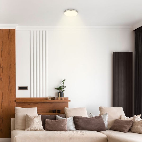 Modernes Wohnzimmer mit einer runden LED-Deckenleuchte, einem beigen Sofa mit Zierkissen und weißen Wänden mit vertikalen Zierleisten.