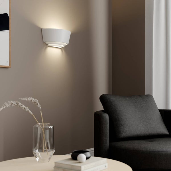 Moderne weiße Wandleuchte mit Stufendesign in einem minimalistischen Wohnzimmer mit dunklem Sessel und Beistelltisch.