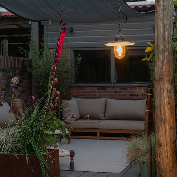Gemütlich beleuchtete Terrasse mit einer Außenpendelleuchte unter einem Sonnensegel, Gartensofa mit Polstern und hohen pinken Blumen.
