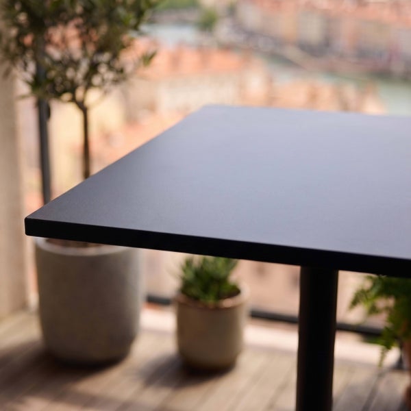Dunkler Outdoor-Tisch mit matter Oberfläche auf einem Balkon mit Topfpflanzen und unscharfem Stadthintergrund.
