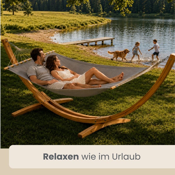 Paar entspannt in grauer Hängematte mit bogenförmigem Holzgestell auf einer Wiese am See, im Hintergrund spielende Kinder und ein Hund.