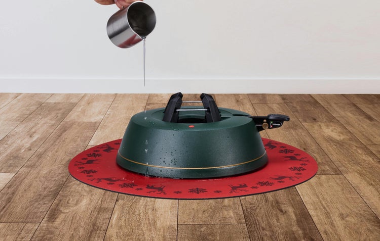 Christbaumständer auf einer roten Unterlage auf Holzboden, Wasser wird aus einer Edelstahlkanne eingefüllt.