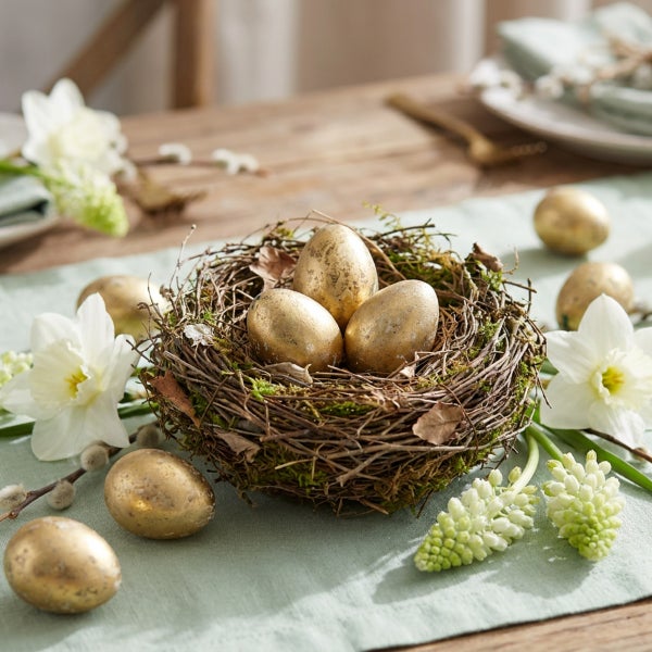 Osternest mit drei goldfarbenen Eiern auf einem Holztisch, dekoriert mit weißen Narzissen und Traubenhyazinthen auf einem hellen Tischläufer.