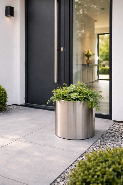 Zylindrischer Pflanzkübel aus Edelstahl mit grünen Pflanzen auf einer modernen Terrasse vor einer Haustür.