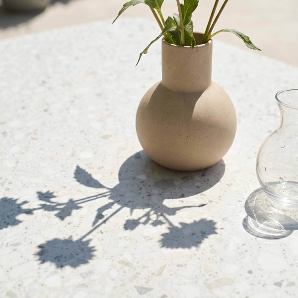 Moderner Terrazzo-Tisch mit runder Keramikvase mit Pflanzenstielen und kleiner Glasvase in heller Umgebung.