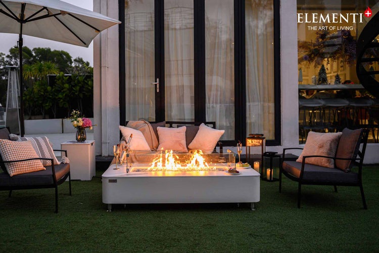 Rechteckiger weißer Gas-Feuertisch mit Glas-Windschutz in einer modernen Gartenlounge mit Polstermöbeln und Abendbeleuchtung. Elementi Logo.