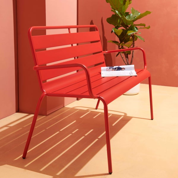 Moderne Gartenbank aus rotem Metall mit Querstreben in einer hellen Umgebung neben einer Zimmerpflanze.