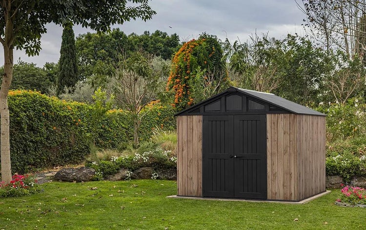 Modernes Gerätehaus in Holzoptik mit dunklen Doppeltüren auf einer Rasenfläche in einem Garten mit üppiger Vegetation.