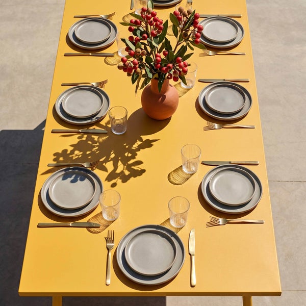 Gedeckter Gartentisch mit gelber Metallplatte, grauen Keramiktellern, goldenem Besteck, Trinkgläsern und Vase mit Beerenzweigen.