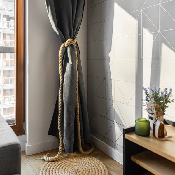 Vorhang mit Seil-Raffhalter, runder Seilteppich auf Holzboden vor einer Wand mit geometrischem Dreiecksmuster. Hell und modern gestaltet.
