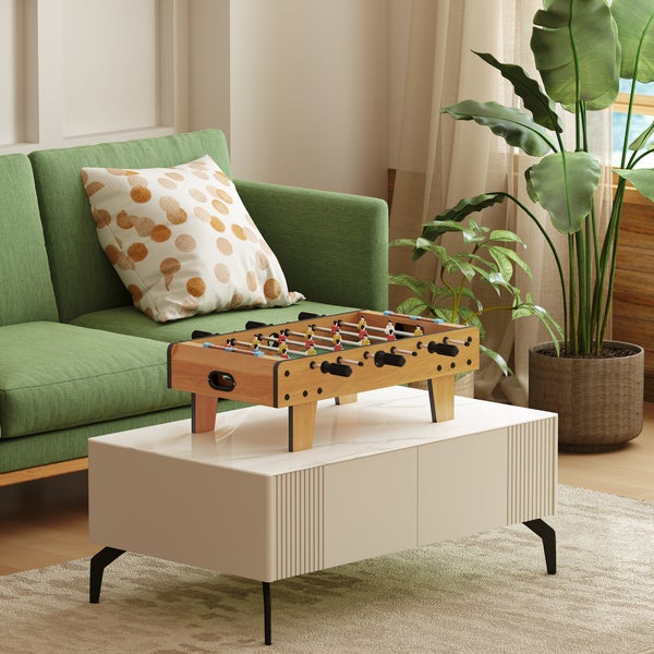 Tischkicker in Holzoptik auf einem weißen Couchtisch in einem hellen Wohnzimmer mit grünem Sofa und großer Zimmerpflanze.