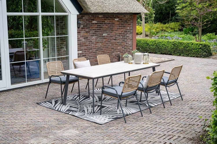 Modernes Gartenmöbel-Set mit rechteckigem Esstisch, sechs Stühlen mit Geflecht-Rückenlehne und einem gemusterten Außenteppich auf einer Steinterrasse.