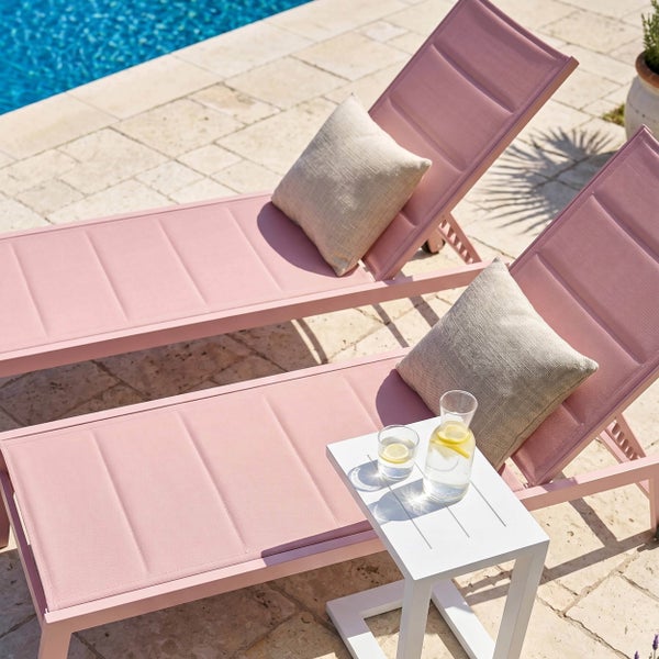 Zwei rosa Gartenliegen mit Kissen auf einer Steinterrasse am Pool neben einem weißen Beistelltisch.