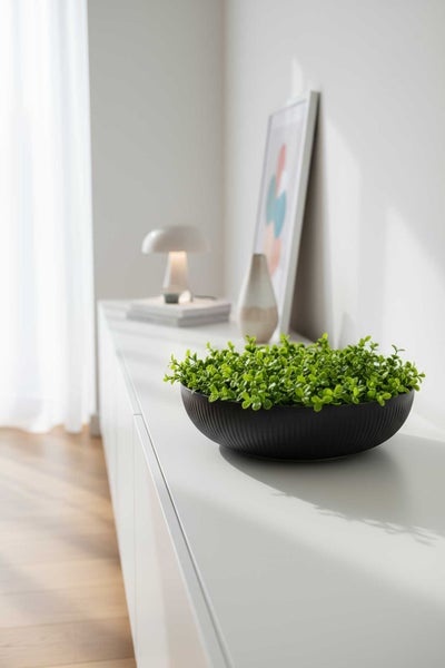 Schwarze runde Dekoschale mit Rillenstruktur und grüner Kunstpflanze auf einem weißen Sideboard in einem hellen modernen Raum.