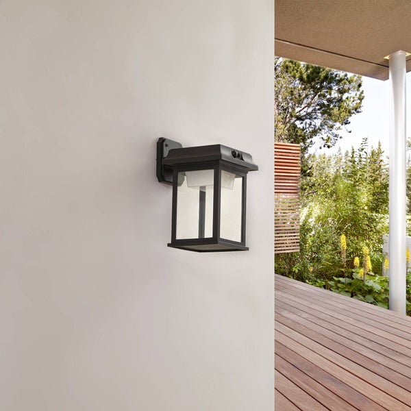 Schwarze Außenwandleuchte im Laternenstil mit Bewegungsmelder an einer Fassade neben einer hellen Holzterrasse mit Gartenblick.