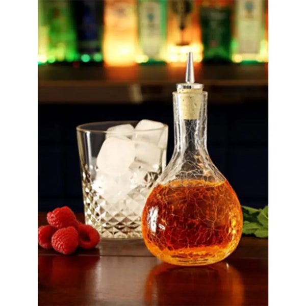 Strukturierte Glaskaraffe mit Metallausgießer auf einer Holztheke neben einem Glas mit Eiswürfeln und Himbeeren.