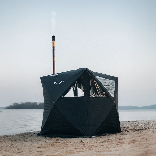 Tragbare Zeltsauna von RUNA auf einem Sandstrand am Wasser mit Schornstein und Fenstern.