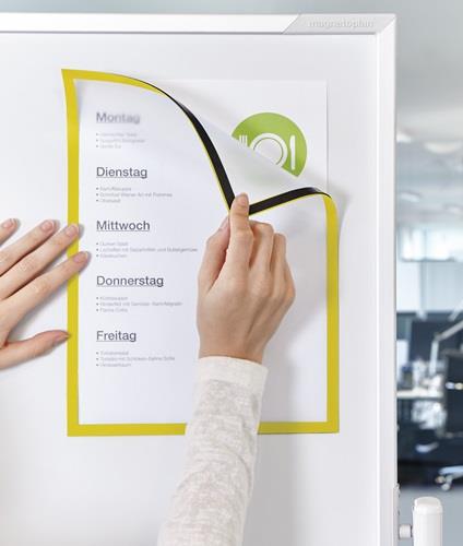 Eine Hand befestigt einen gelben Magnetrahmen mit einem Speiseplan an einer Whiteboard-Oberfläche