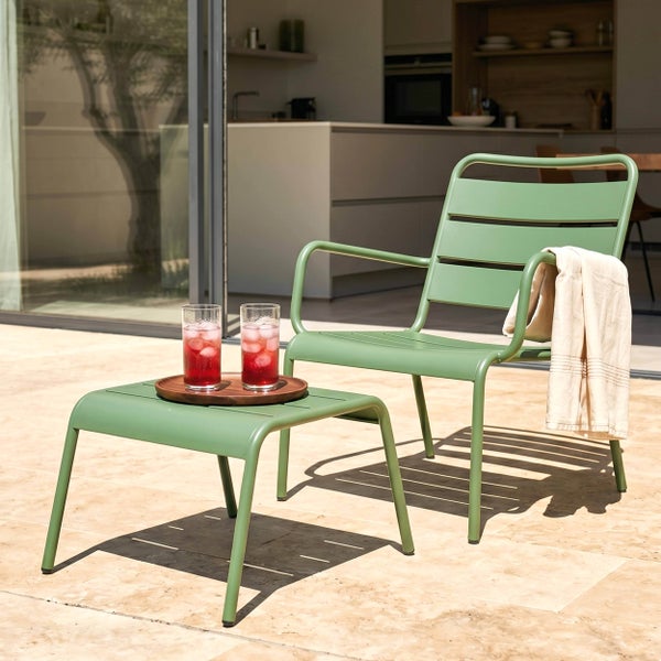 Moderne Garten-Loungegarnitur aus grünem Metall bestehend aus Sessel und Hocker auf einer hellen Steinterrasse in sonniger Atmosphäre.