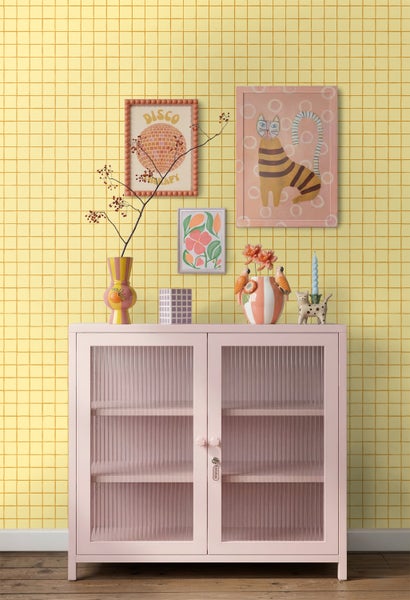 Rosa Metallvitrine mit Riffelglas vor einer hellgelben Tapete mit Karomuster. Dekorative Vasen und gerahmte Bilder im Retro-Stil.